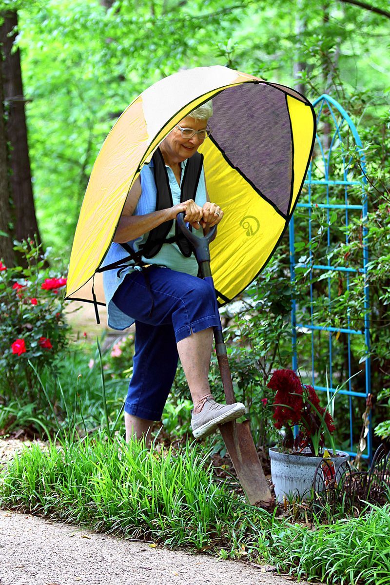 Woman wearing the GOShelter Shade sun protection while gardening. Stands on a wooden plank step with flowers in a garden.