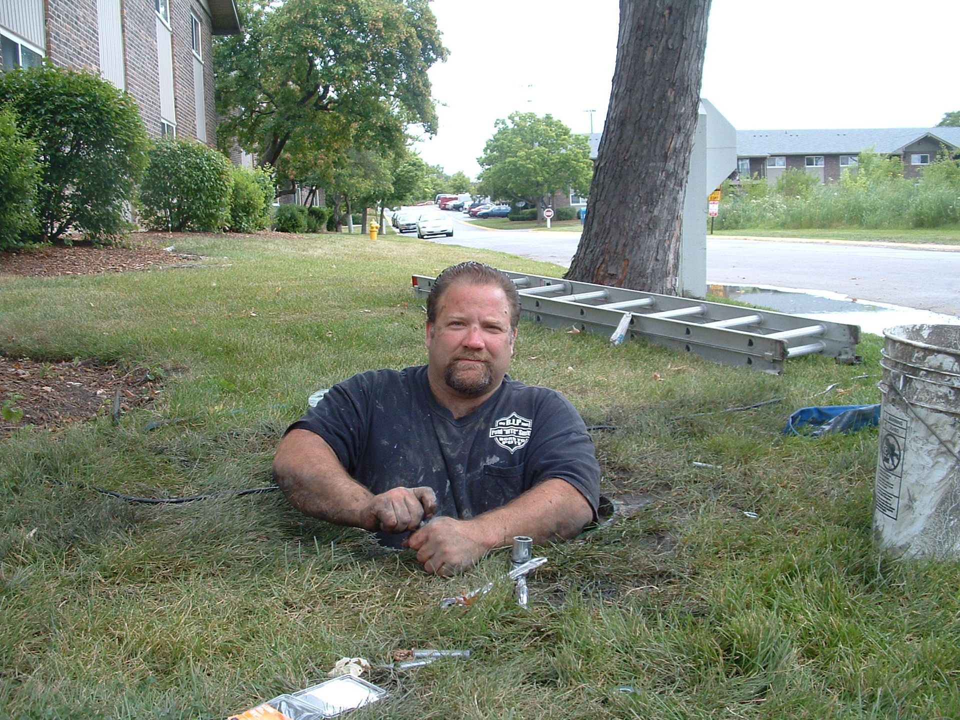 Man in a hole in the ground, looking up. He's on a grassy lawn near a road and buildings.
