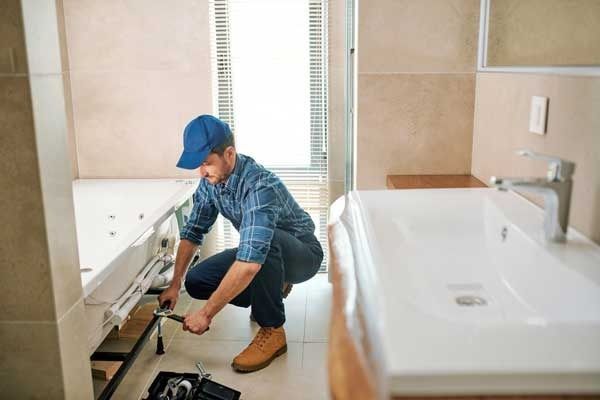 A man in a blue cap and shirt works on plumbing under a white bathtub in a bathroom.