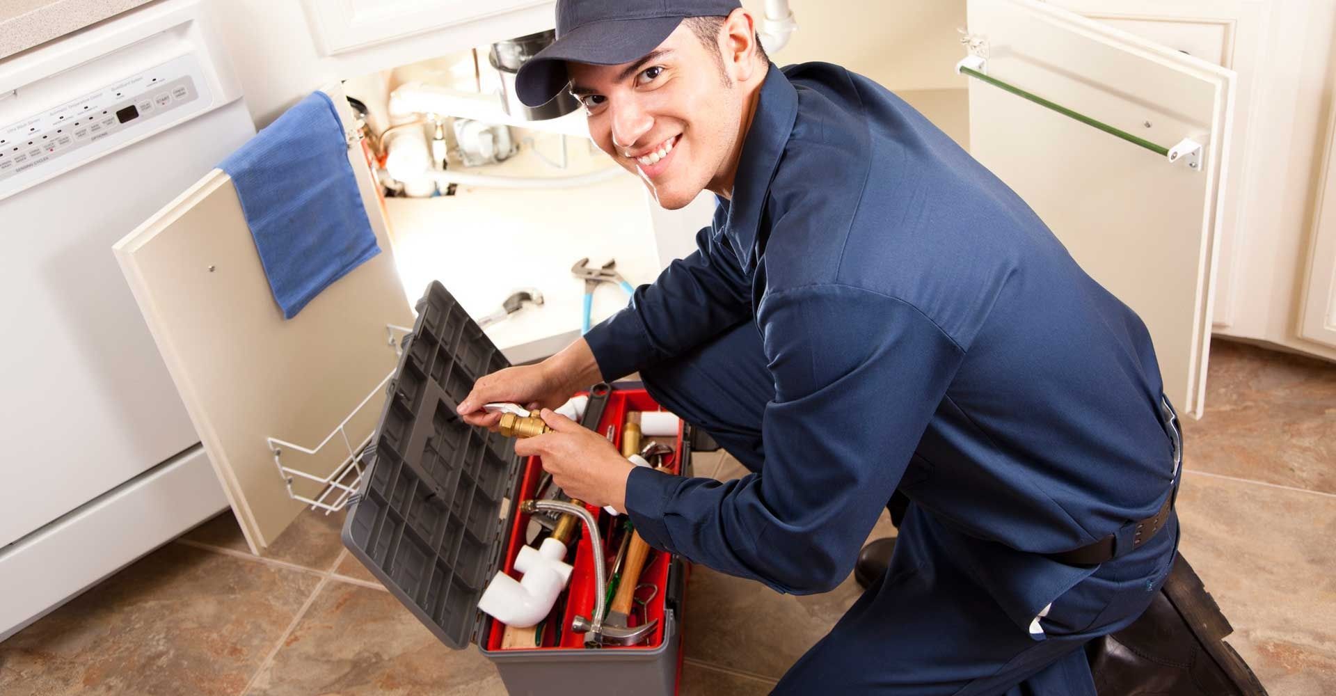Plumber in blue coveralls and hat kneeling with toolbox under sink, smiling.