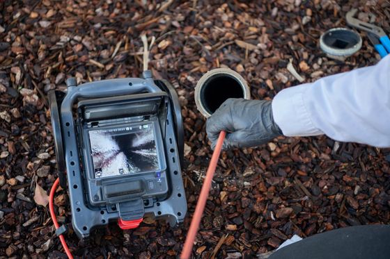 A gloved hand feeds a sewer camera cable into an open pipe while the display shows the internal view of the drain.