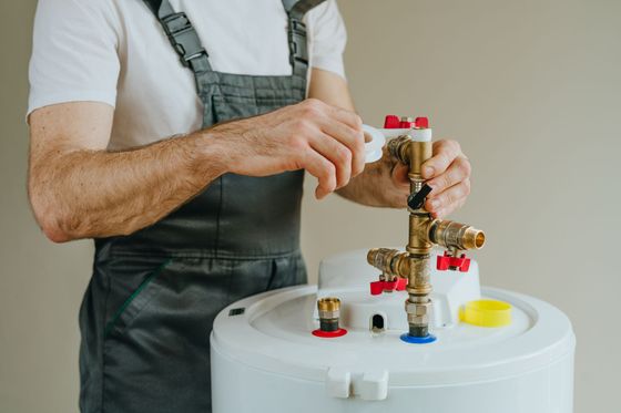 A plumber in gray overalls applies white plumbing tape to the threaded fitting of a water heater assembly.