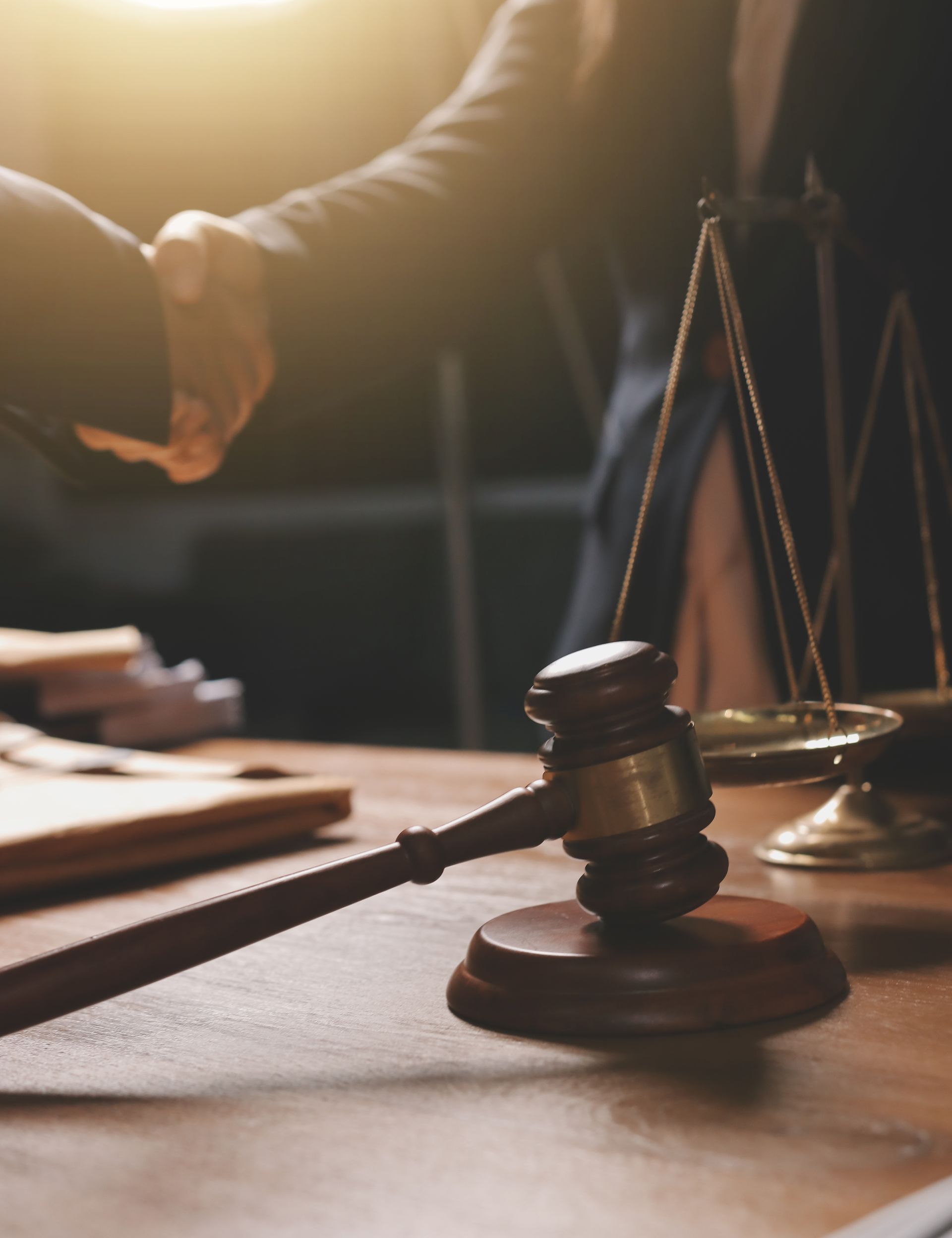 Handshake beside gavel and scales of justice on a wooden desk in warm courtroom light