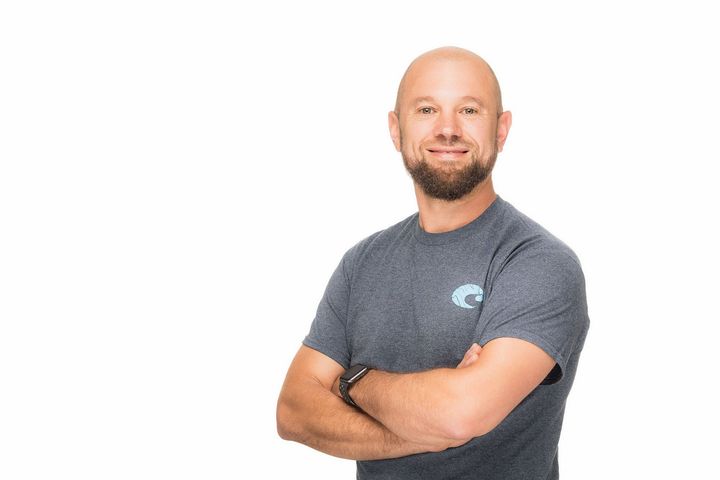 A bald man with a beard is standing with his arms crossed and smiling.