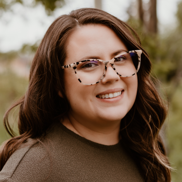 Woman with brown hair and glasses smiles outdoors.