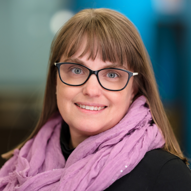 Woman with glasses and pink scarf smiles at the camera.