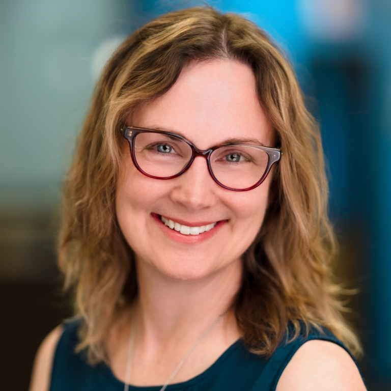 Woman with glasses smiles; blonde hair, teal top, blurred blue background.