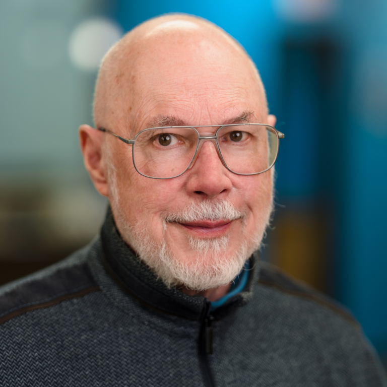 Bald man with glasses and gray beard smiles, wearing a gray and black zipped jacket; blurred blue background.