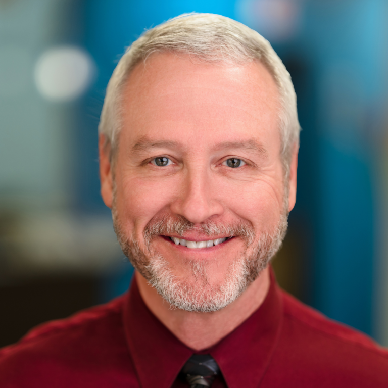 Smiling man with gray hair and beard, wearing a burgundy shirt and black tie, set against a blurred blue background.