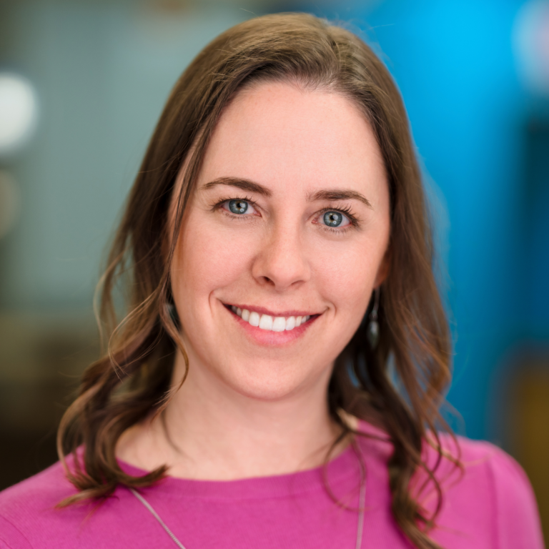 Woman with brown hair smiles, wearing a pink shirt, in front of a blue background.