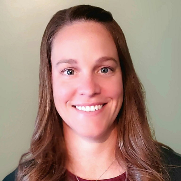 Woman with long brown hair smiling at the camera, wearing a dark top, against a pale green background.