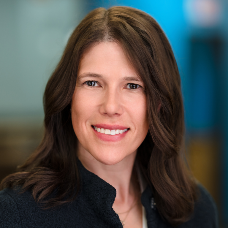 Woman with brown hair smiles, wearing a dark jacket, against a blurred blue background.