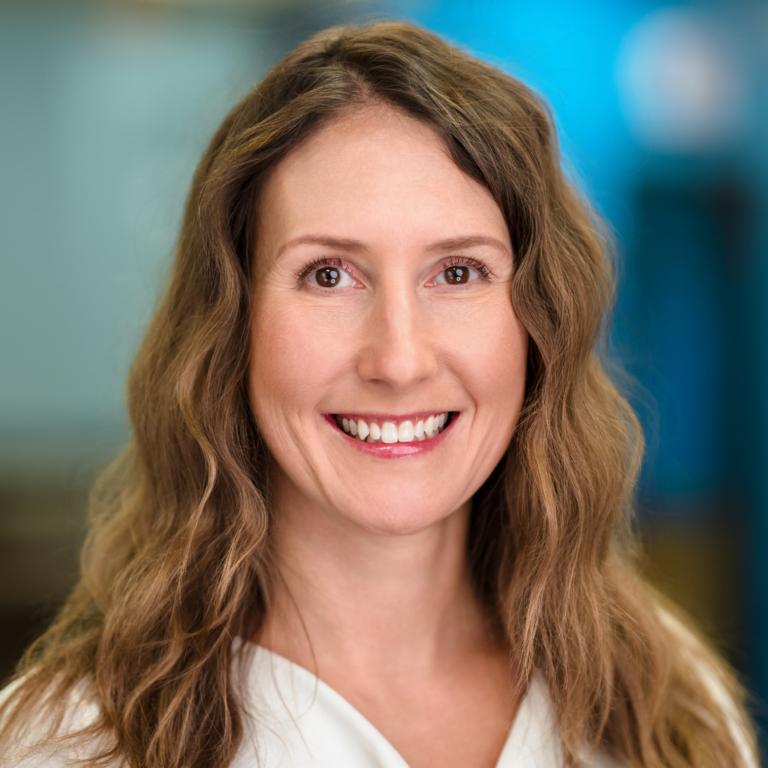 Woman with long wavy brown hair smiles at the camera, blurred blue background.