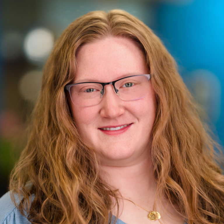 Woman with long, wavy auburn hair, glasses, smiling, wearing blue shirt, gold necklace; blurred background.