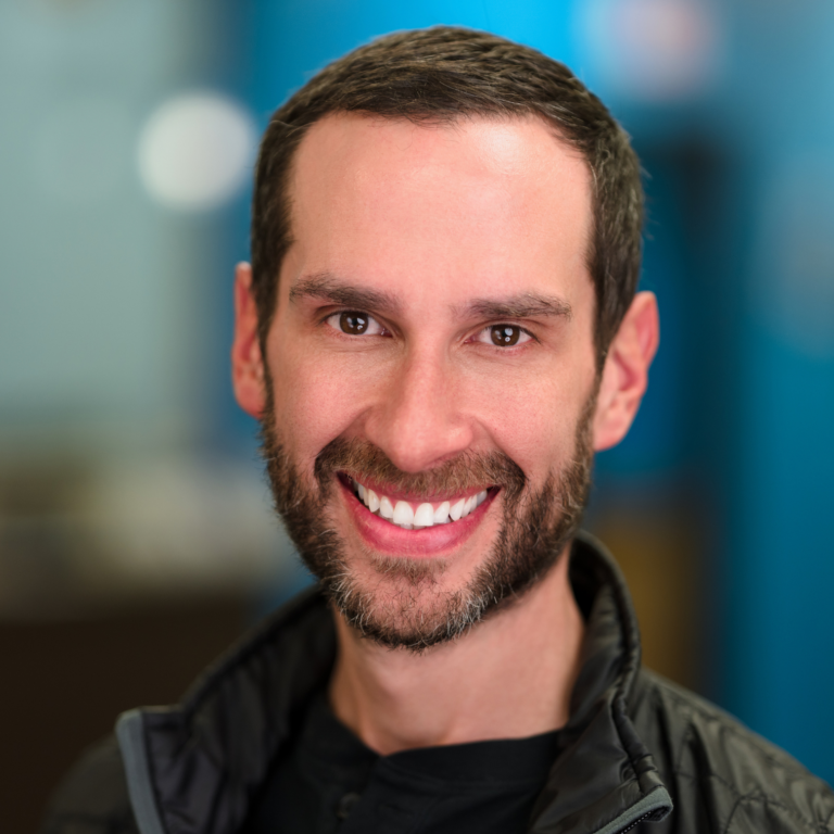 Man with brown hair and beard smiles broadly in front of a blue backdrop. He wears a black shirt and a jacket.