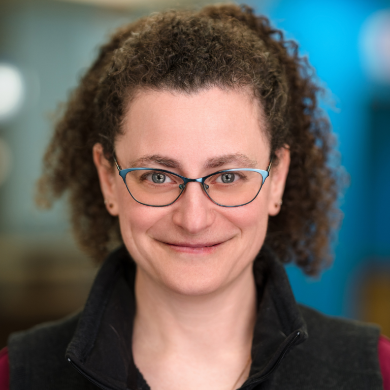 Woman with curly hair and glasses smiles at the camera, wearing a black vest against a blurred blue background.
