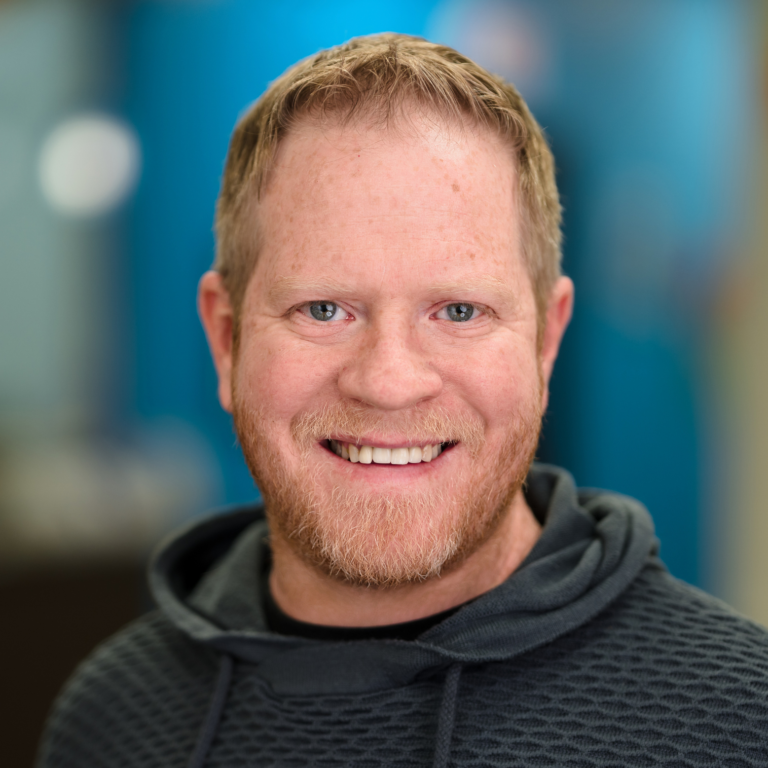 Man with red hair and beard smiling, wearing a gray hoodie, blue background.