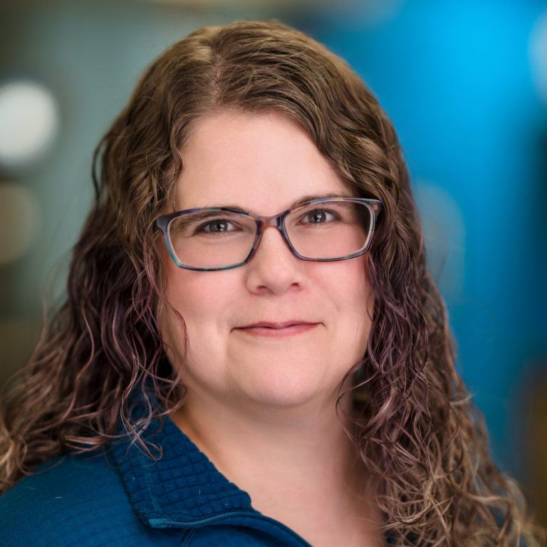 Woman with curly brown hair, glasses, and a blue shirt smiles against a blurred blue background.