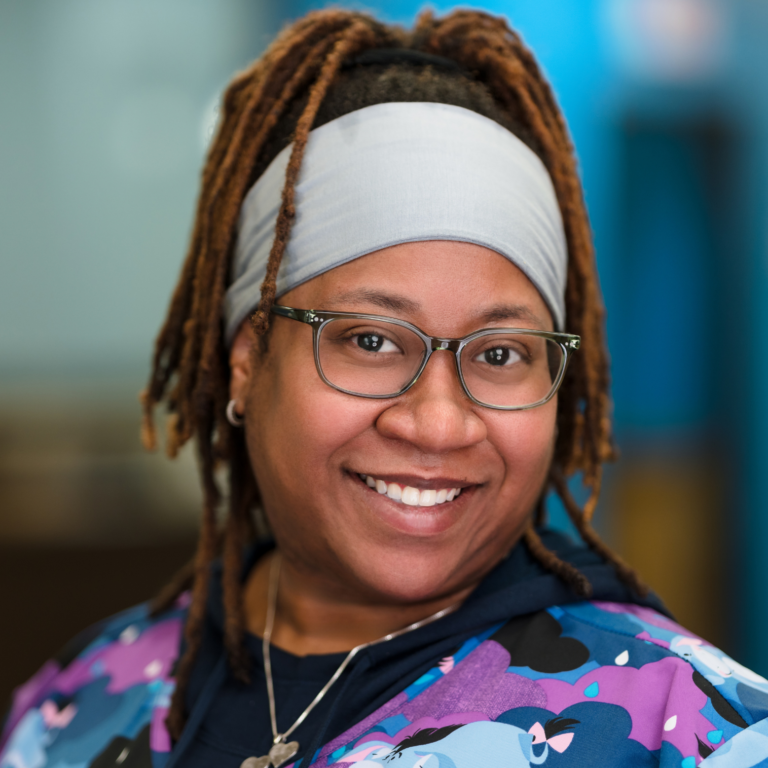 Woman with dreadlocks, wearing glasses, smiling. Wearing a gray headband and patterned jacket.
