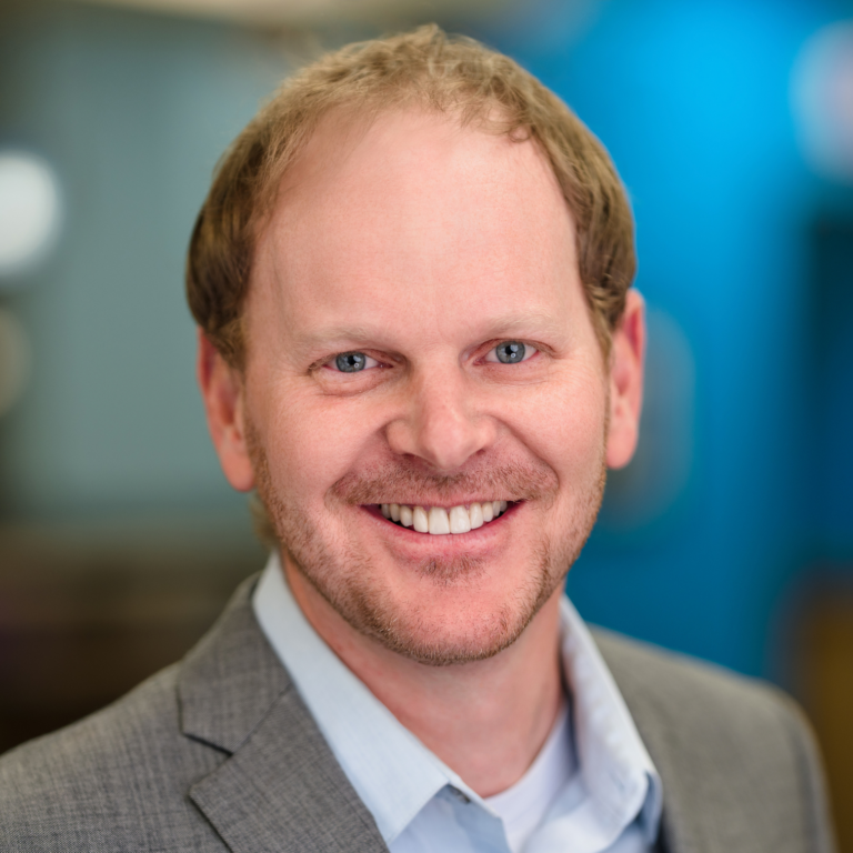 Man in gray blazer smiling at the camera, blue eyes, blurred background.