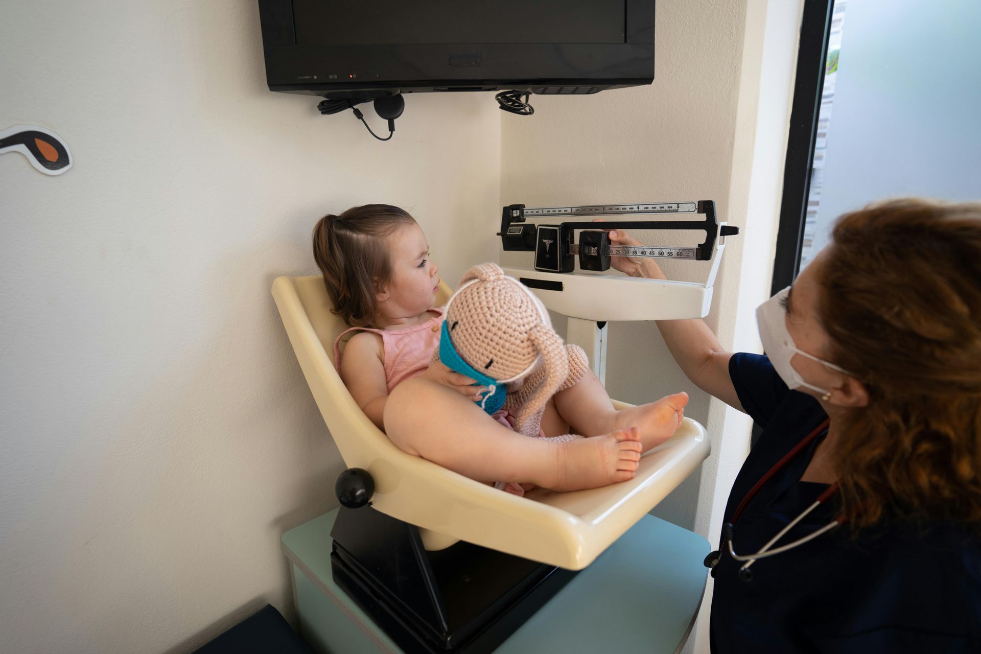 Child sits on scale while a person in scrubs adjusts it indoors.