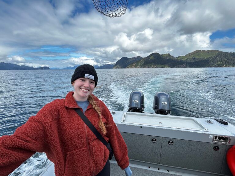 Woman smiles aboard a boat, ocean and mountains in the background, under a cloudy sky.
