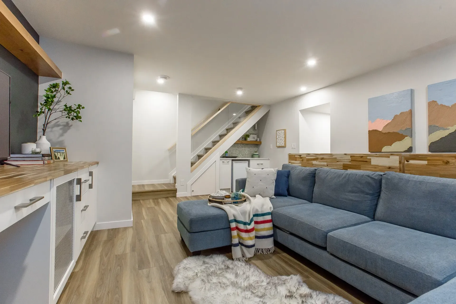 Cozy basement living room with blue sectional, bar, artwork, and stairs leading up.