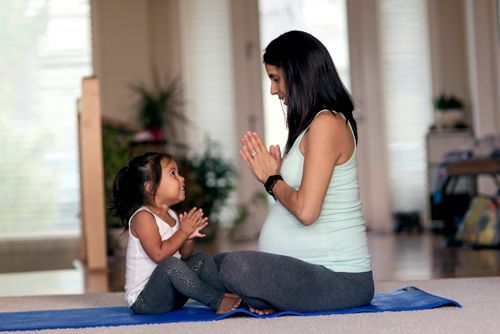 Pregnant woman and child sitting on a yoga mat with hands together, indoors.