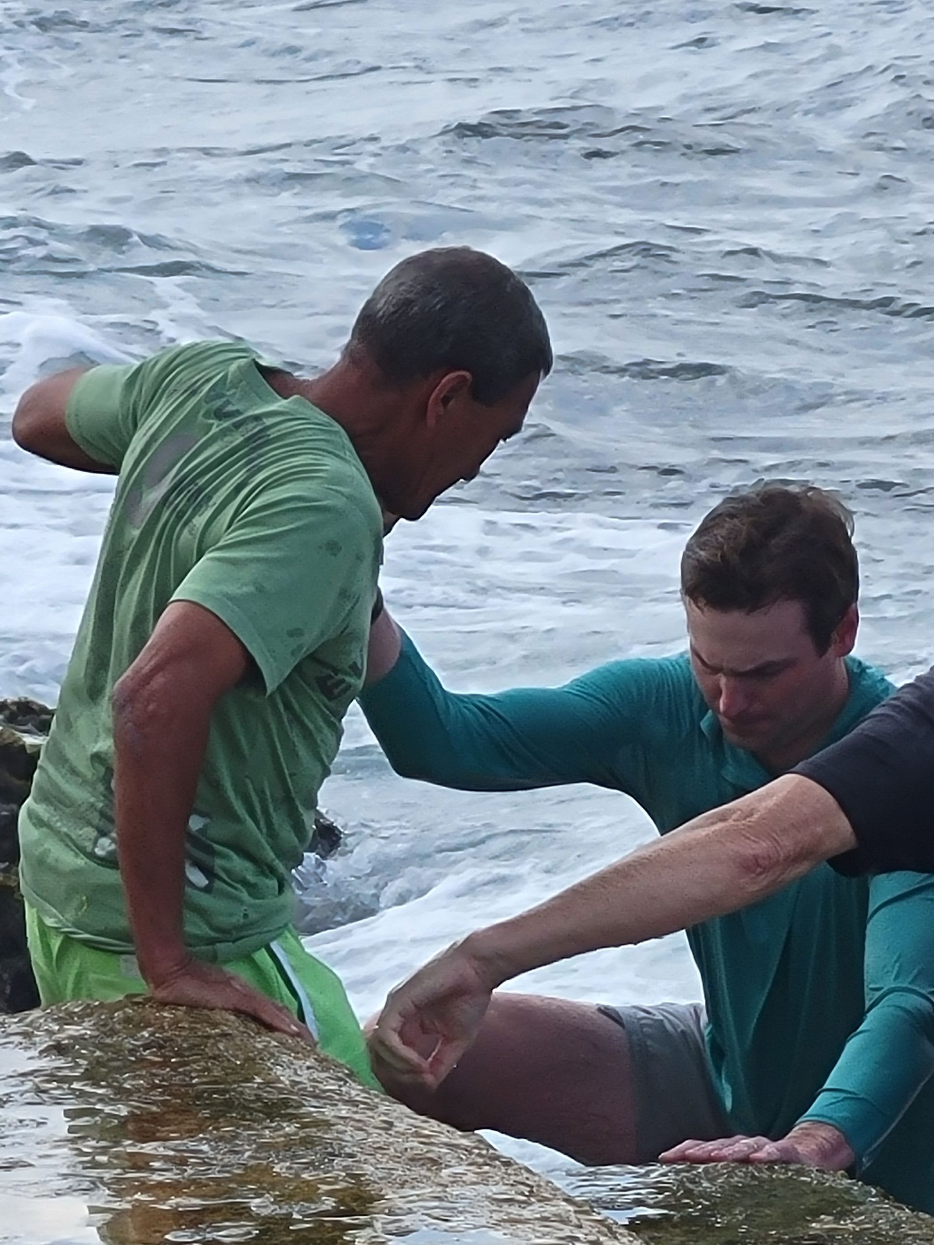 A man in a green shirt is helping another man out of the water