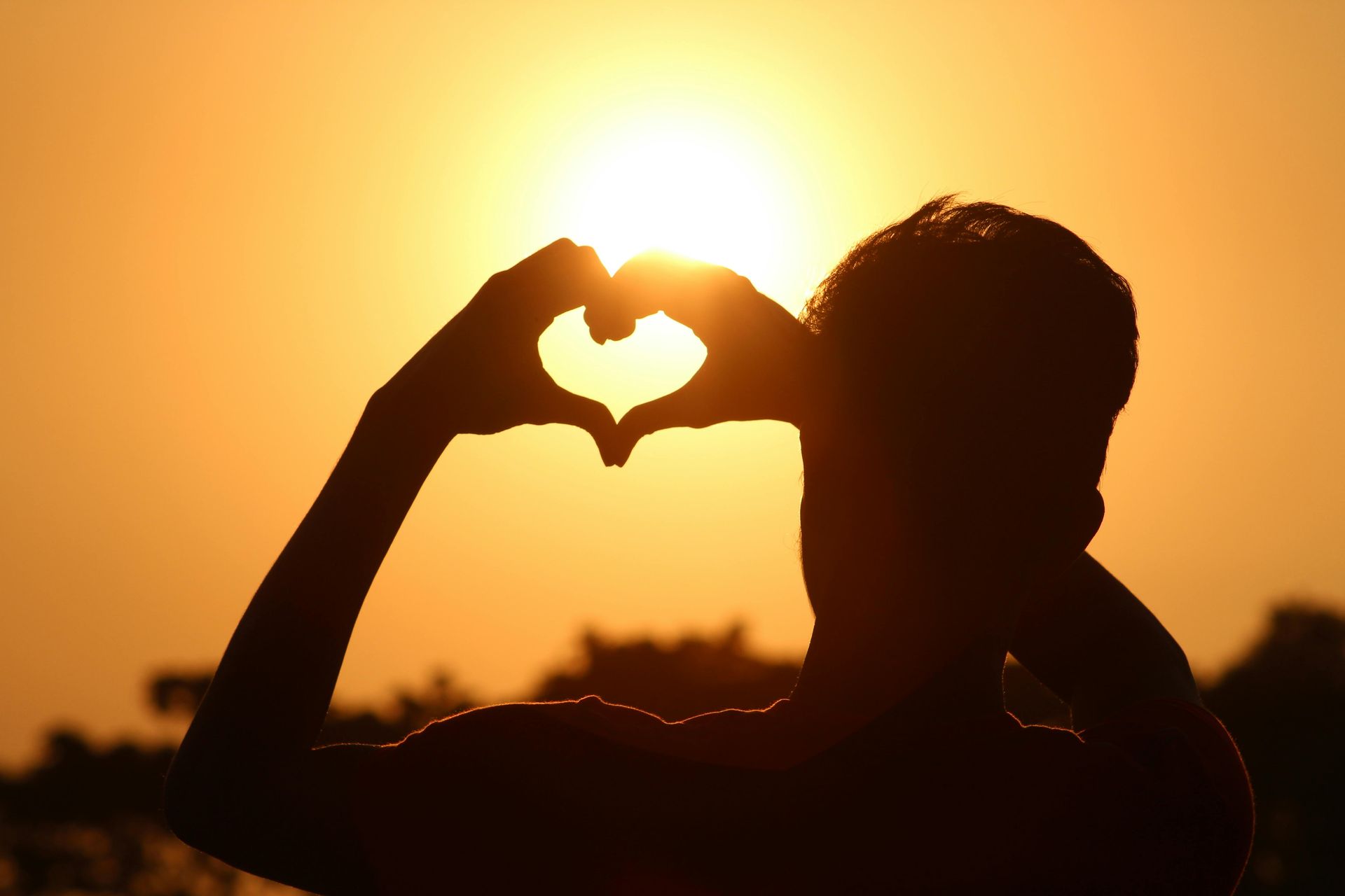 A person making a heart shape with their hands in front of the sun