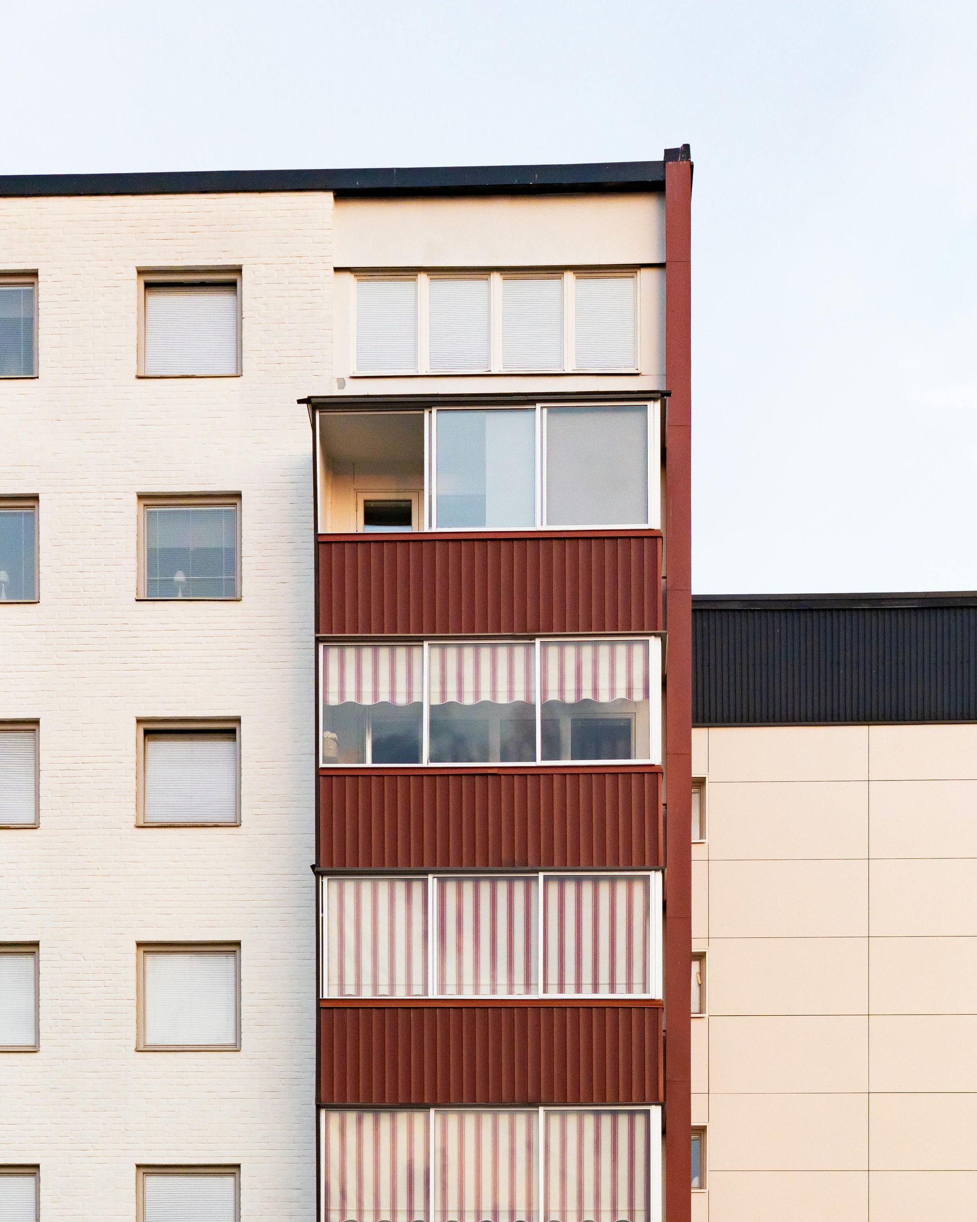 Apartment building facade with red balcony, light-colored brick exterior, and dark roof against a pale sky.