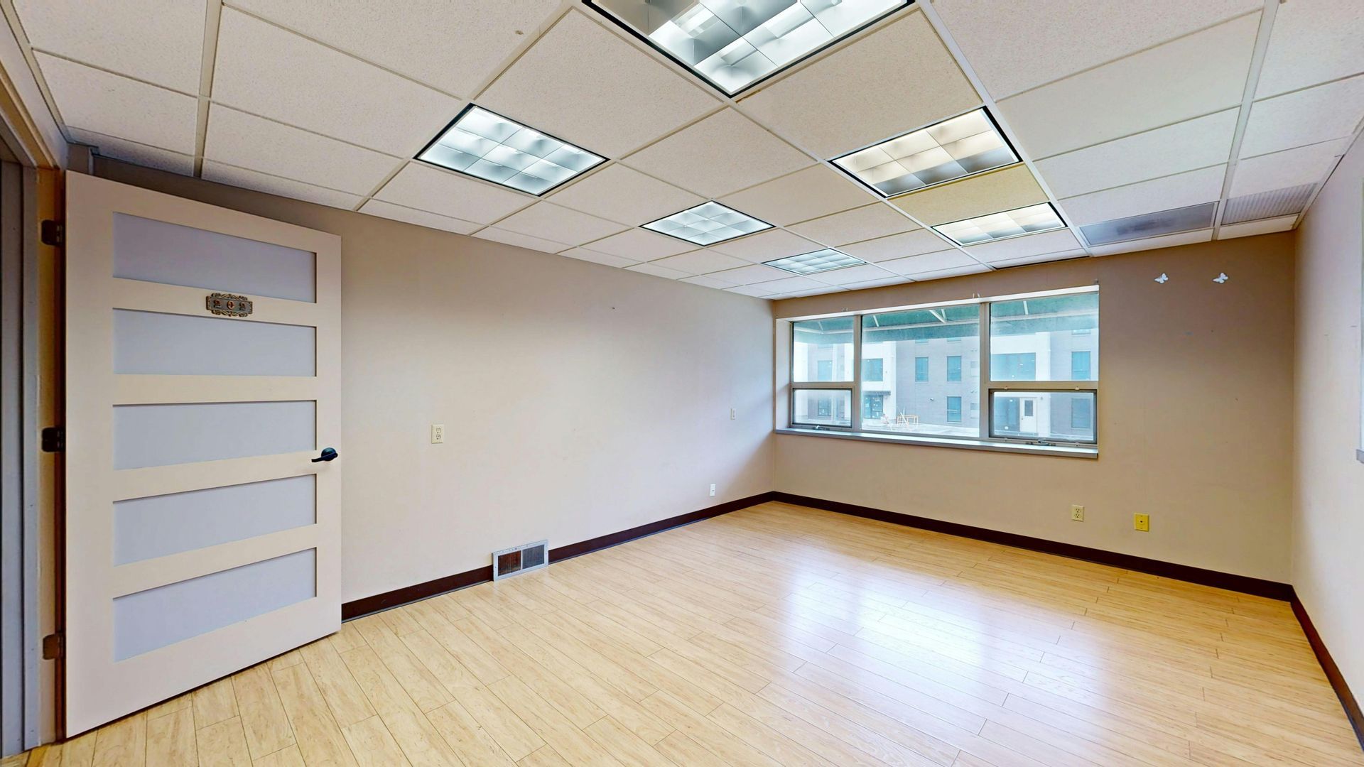 Empty office room with wood floor, beige walls, and a window. Fluorescent lights in the ceiling. Door on left.
