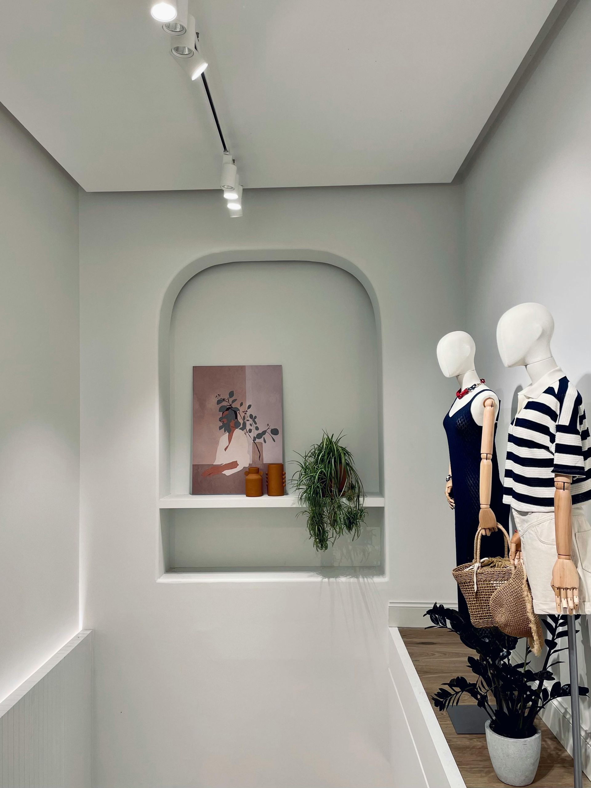 White retail interior with mannequins, arched shelf with art and plant, track lighting.