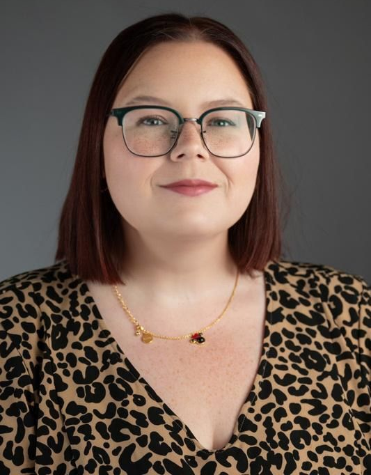 Portrait of a person with glasses and a leopard-print top, wearing a necklace against a gray background