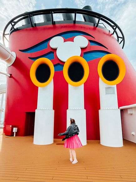 Person in pink skirt stands by a red building with three yellow circular windows and a Mickey Mouse sign.