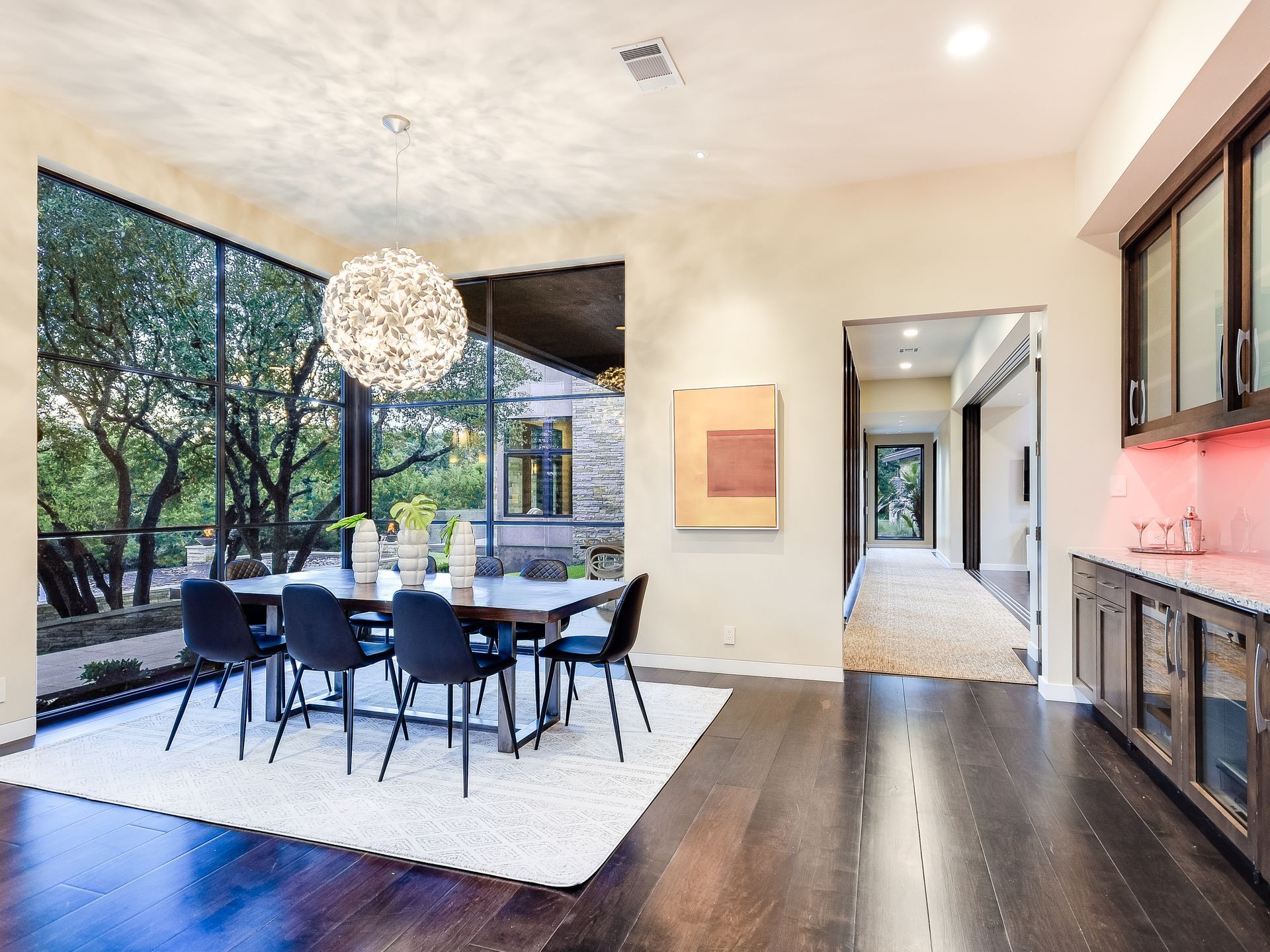 A grand luxury living room with dramatic glass windows overlooking a lush garden, featuring a dining  table, and chandelier