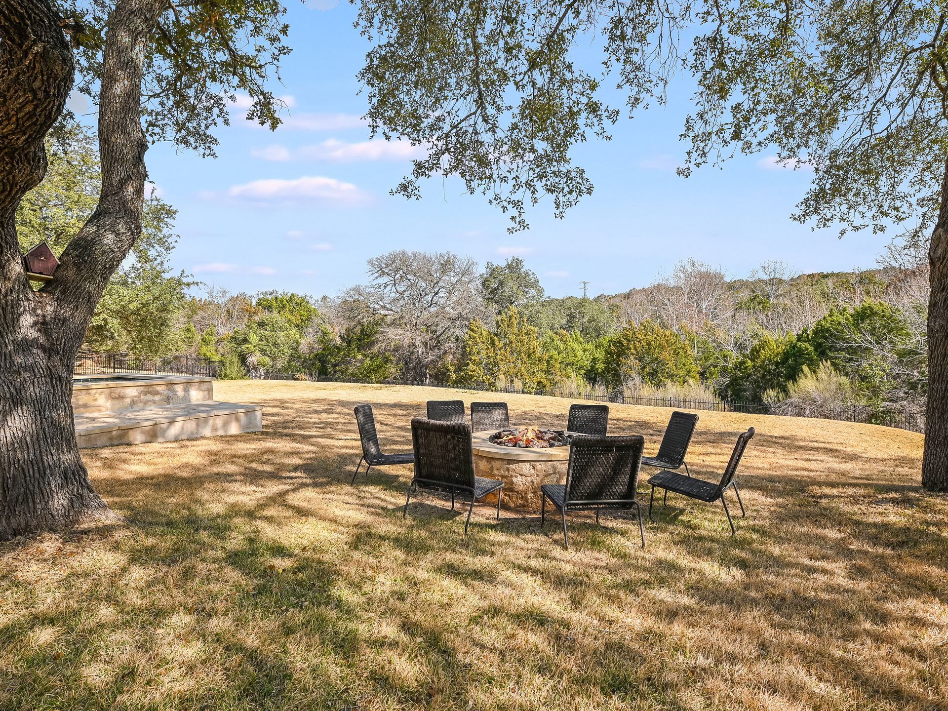 A spacious backyard with a circular firepit surrounded by chairs and outdoor lighting, featuring lush green grass and mature trees.