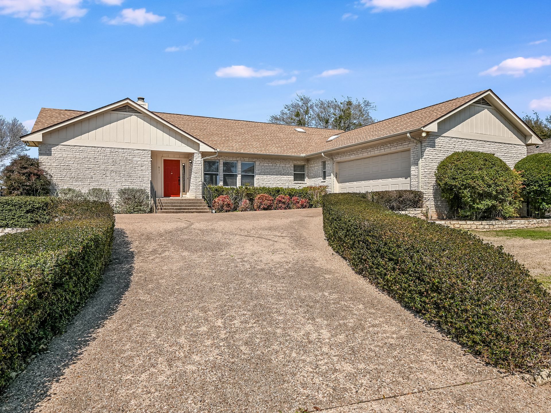 A charming front exterior of a home with a vibrant red door, attached garage, and a beautifully landscaped garden with flowering plants and shrubs