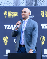 A man stands at a podium with a microphone in front of a sign that says the hidden genius project