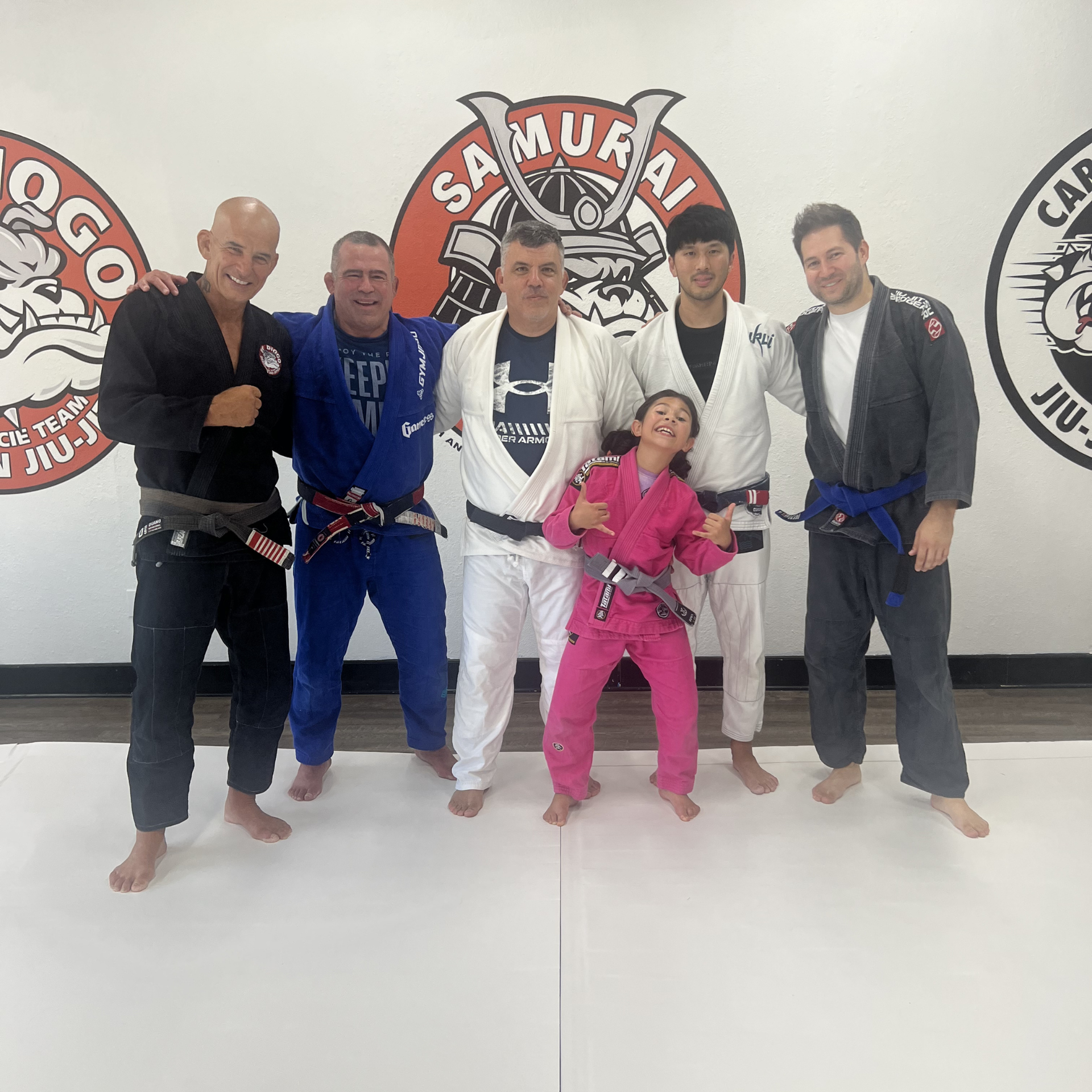 Six people in Jiu-Jitsu gis pose in a line against the Samurai Brazilian Jiu-Jitsu gym wall