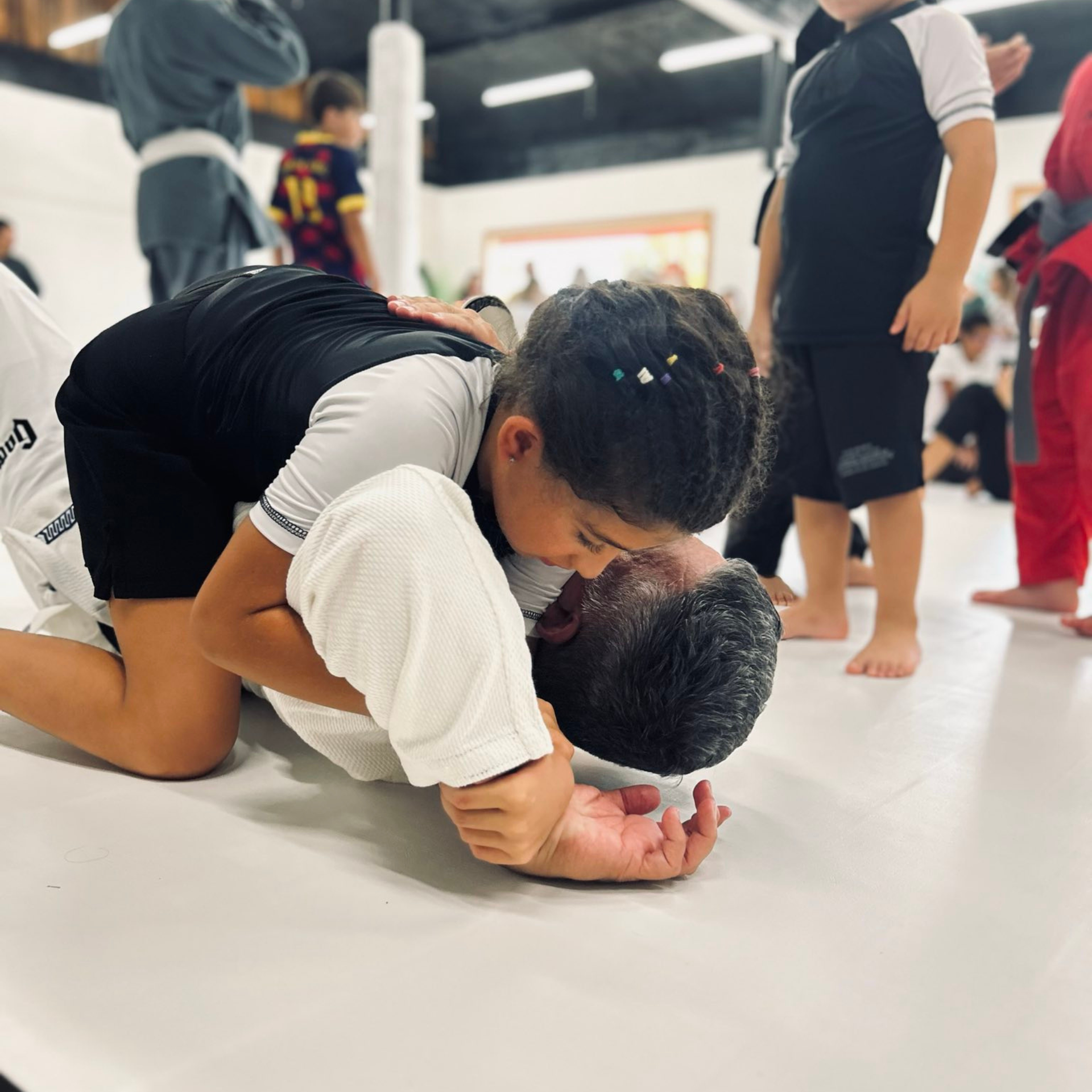 A child wearing a black-and-white rash guard practices a Jiu-Jitsu move on an adult wearing a white gi in a gym.