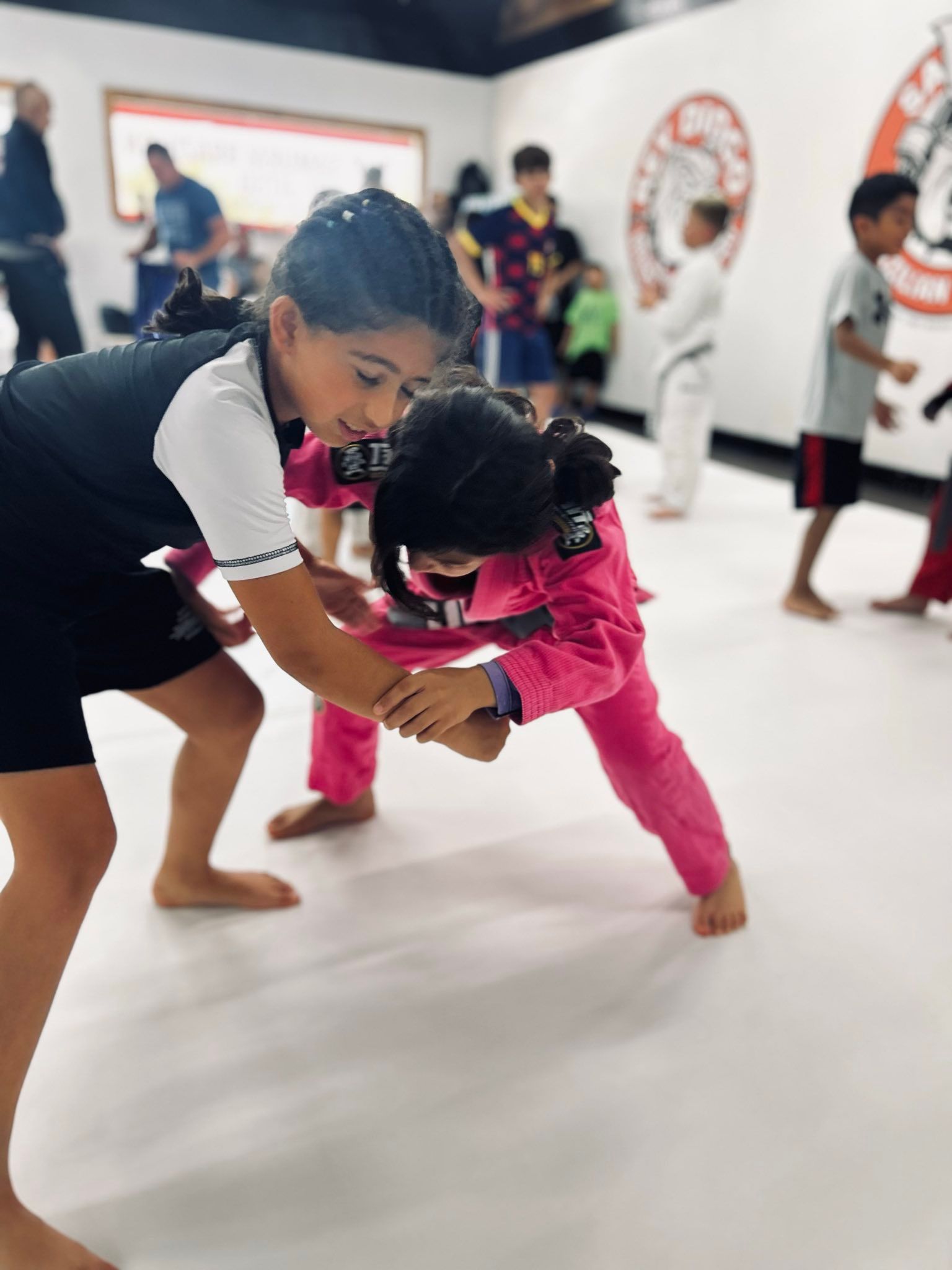 Two children in martial arts uniforms practice a grappling move on a white mat in a training studio.