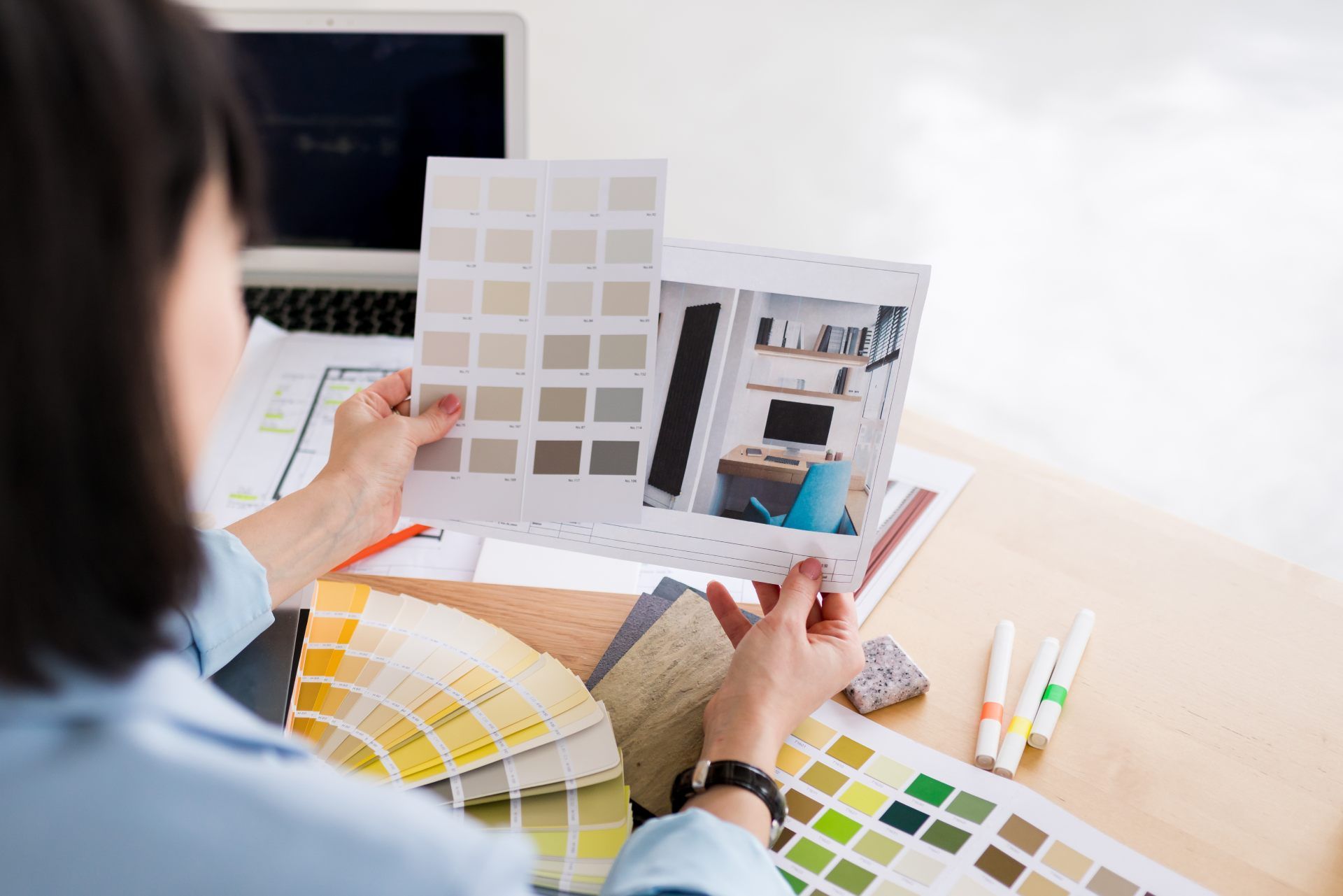 An interior designer reviews color swatches and room renderings on a desk, with paint fans and pens nearby.