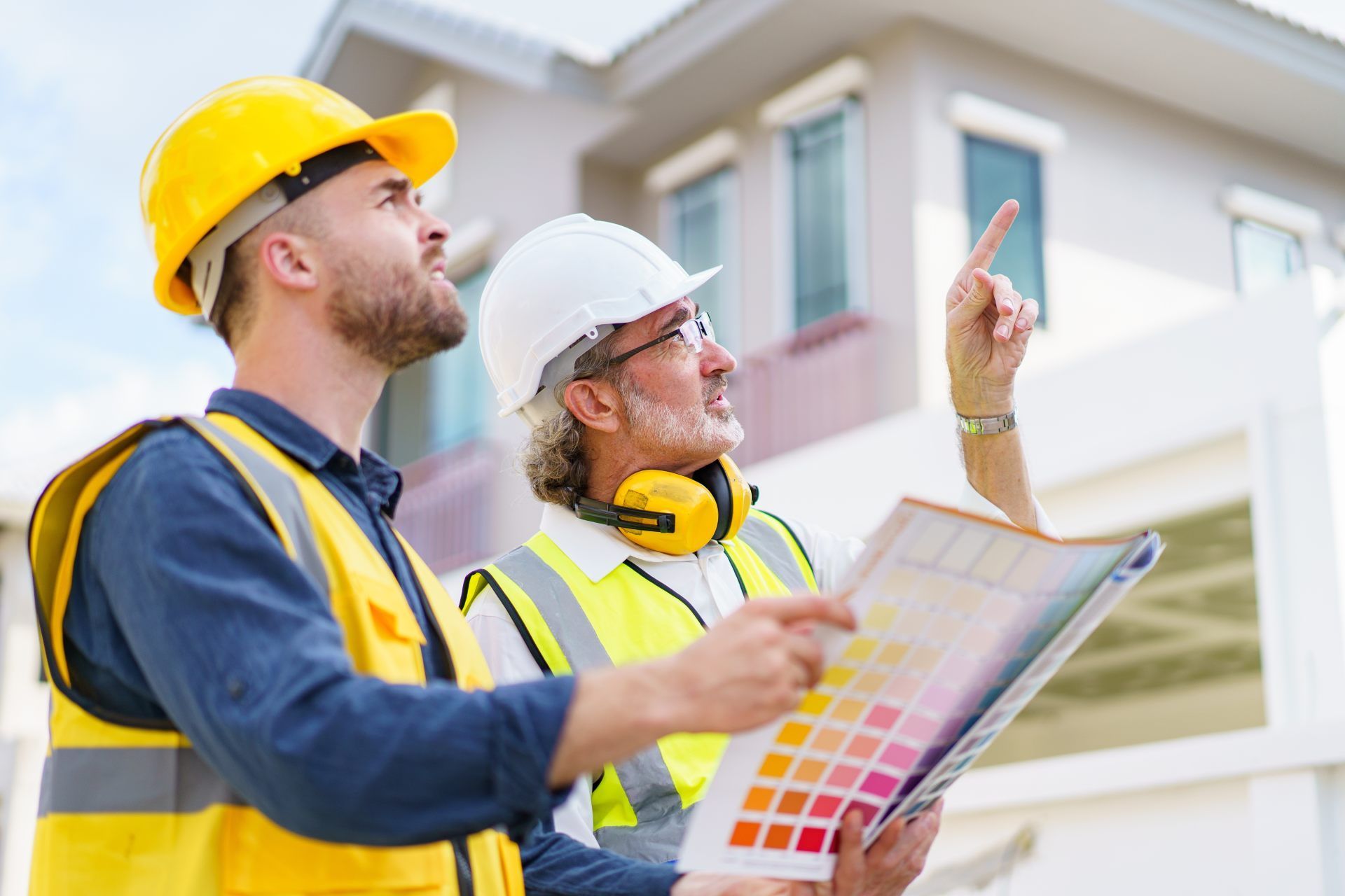 Two construction professionals in high-visibility vests and hard hats look at a paint color swatch while pointing at a house.
