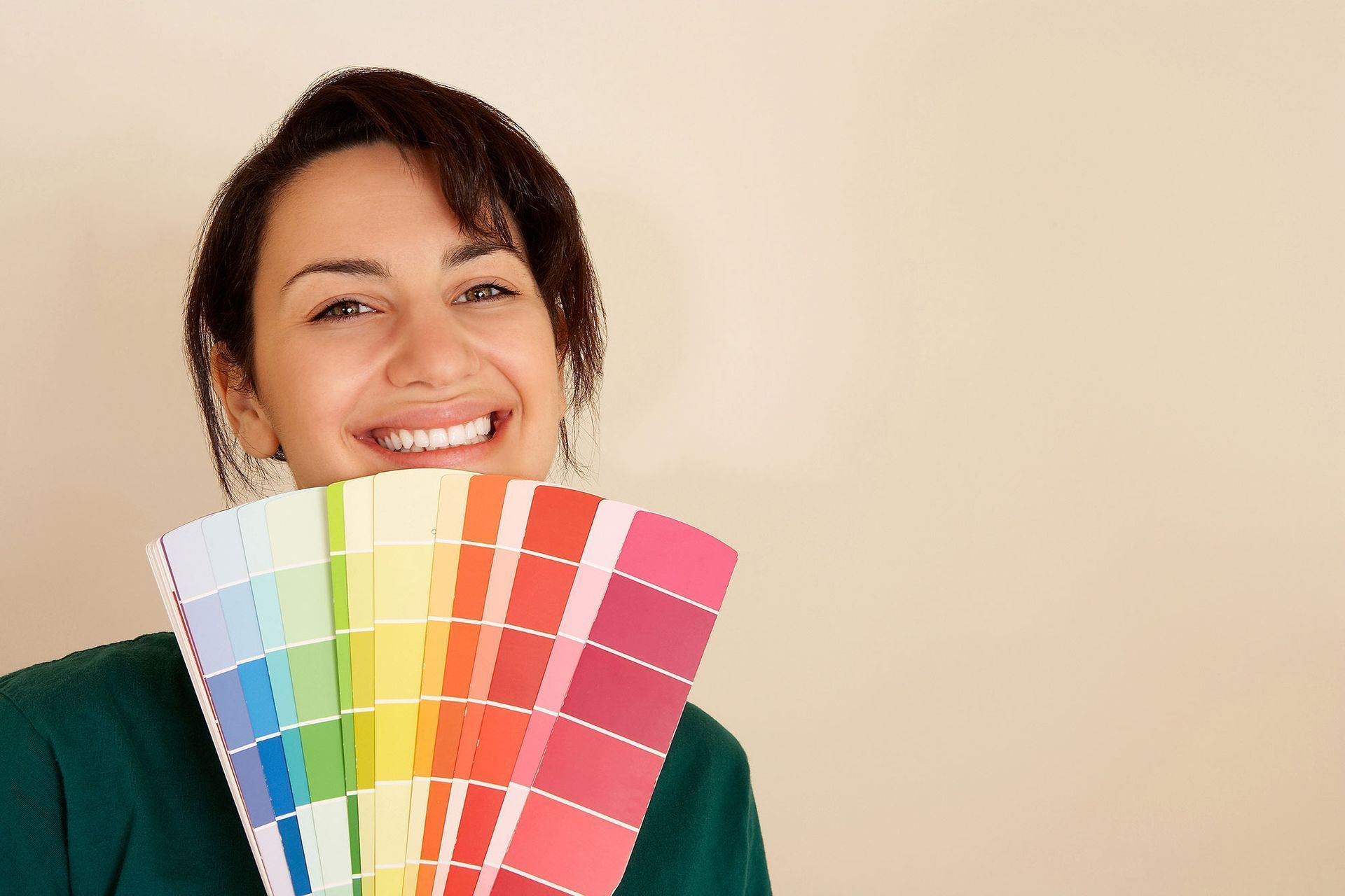 A smiling person holds a multi-colored paint swatch fan in front of their face against a neutral background.