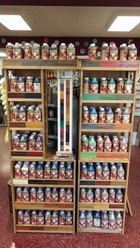 Two wooden display shelves in a store stocked with rows of various paint or stain bottles with color-coded labels.