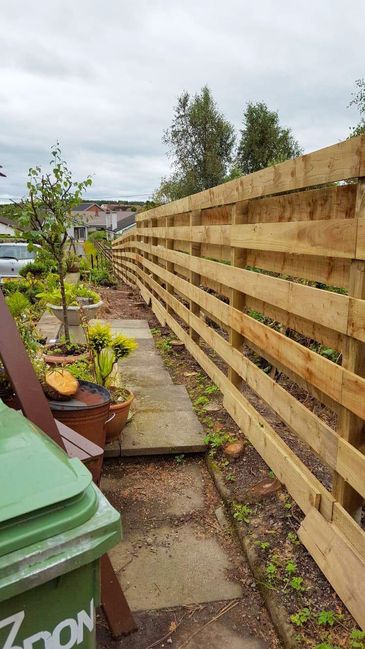 A wooden fence along a sidewalk next to a green trash can.