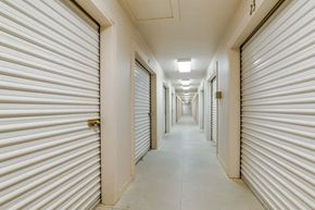 Storage units in a row, with white doors, brick accents, and a concrete drive. Clear blue sky.