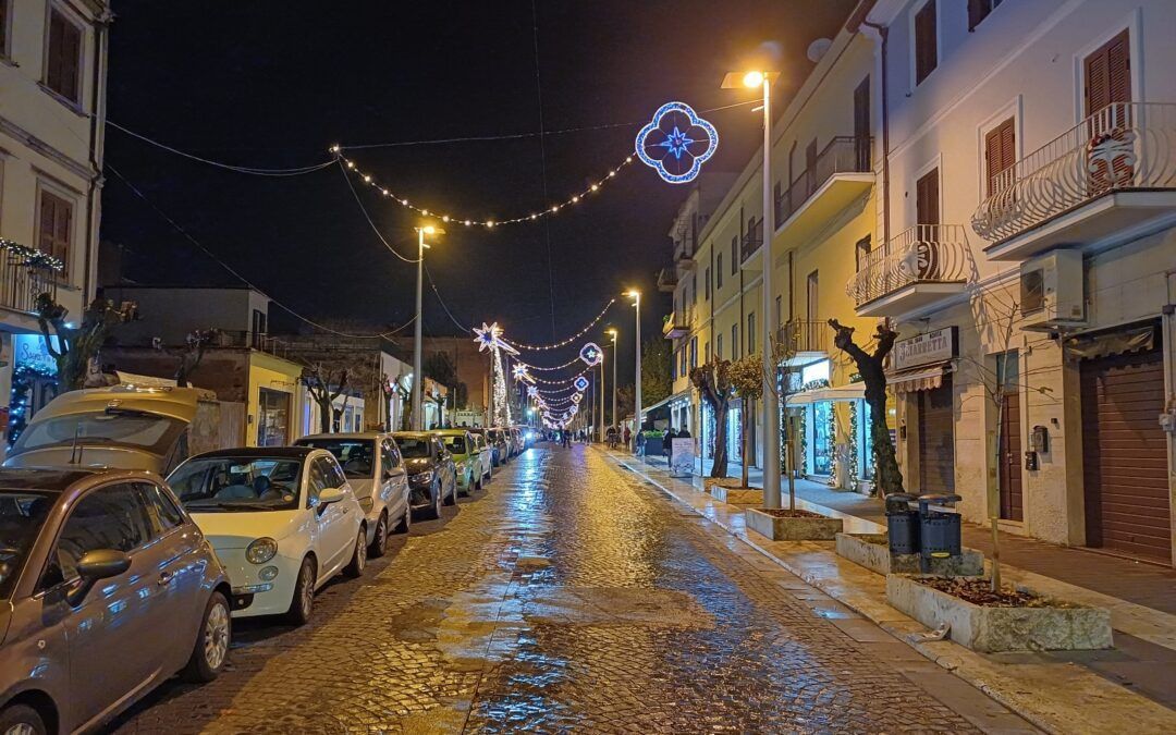 Una veduta notturna di una strada acciottolata fiancheggiata da auto parcheggiate, illuminata da lampioni e decorazioni festive sospese.