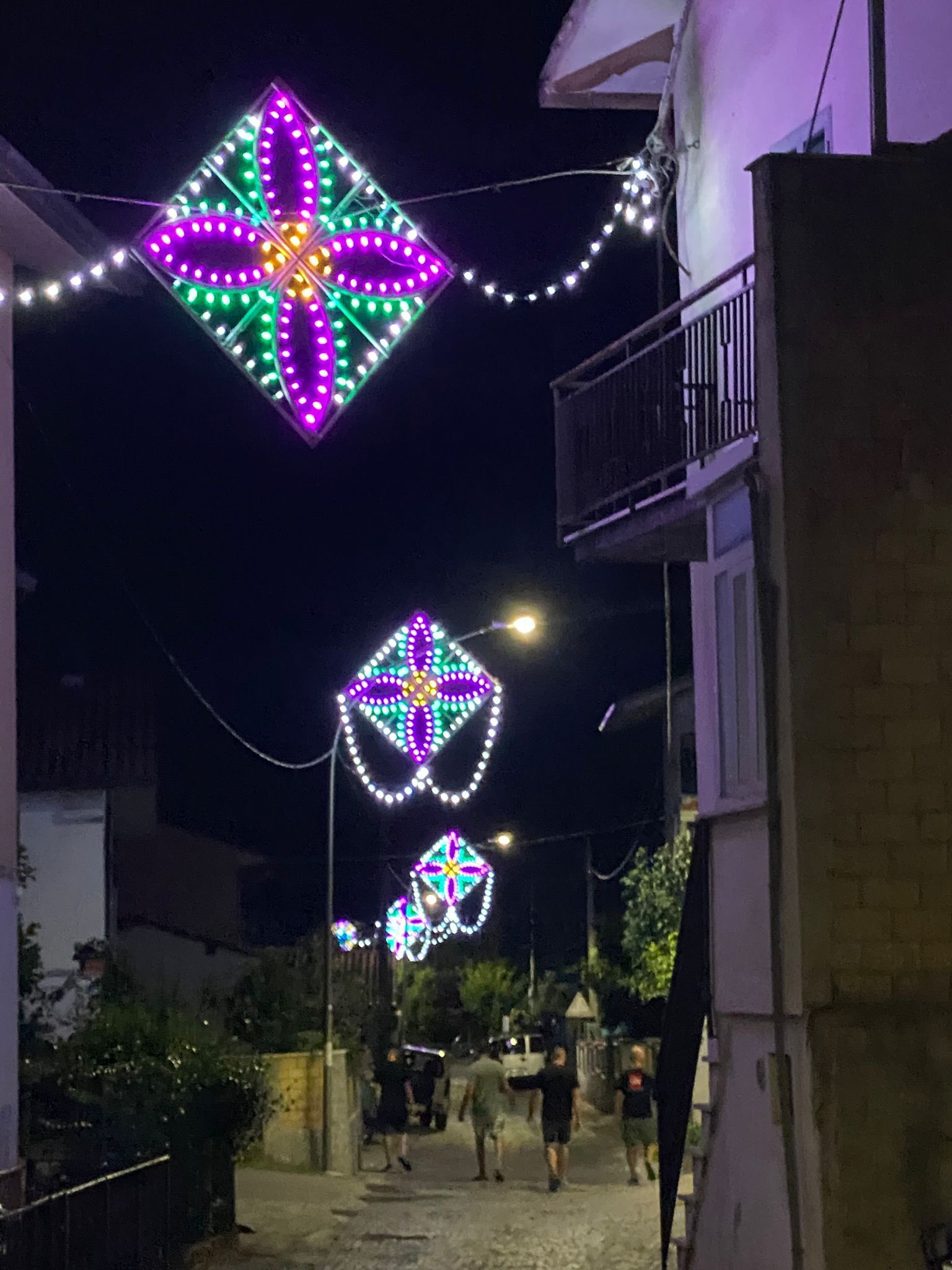 Una strada stretta di notte illuminata da luci decorative a forma di diamante, di colore viola e verde, sospese in alto.